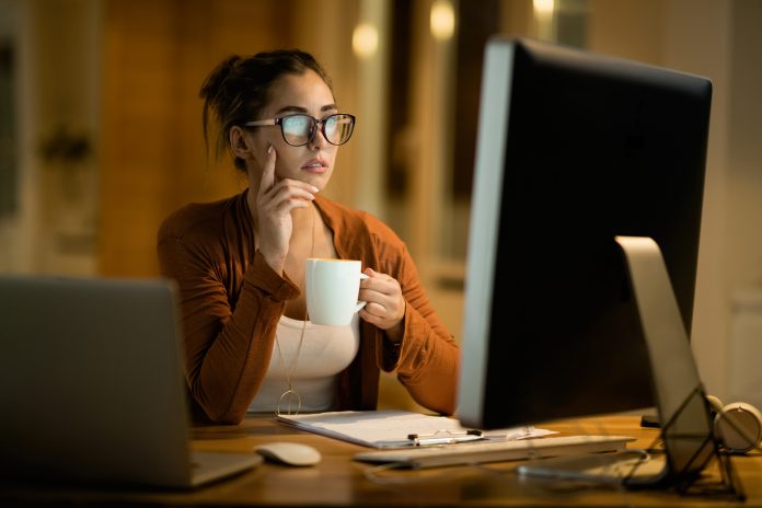 young woman drinking coffee thinking while working desktop pc evening home